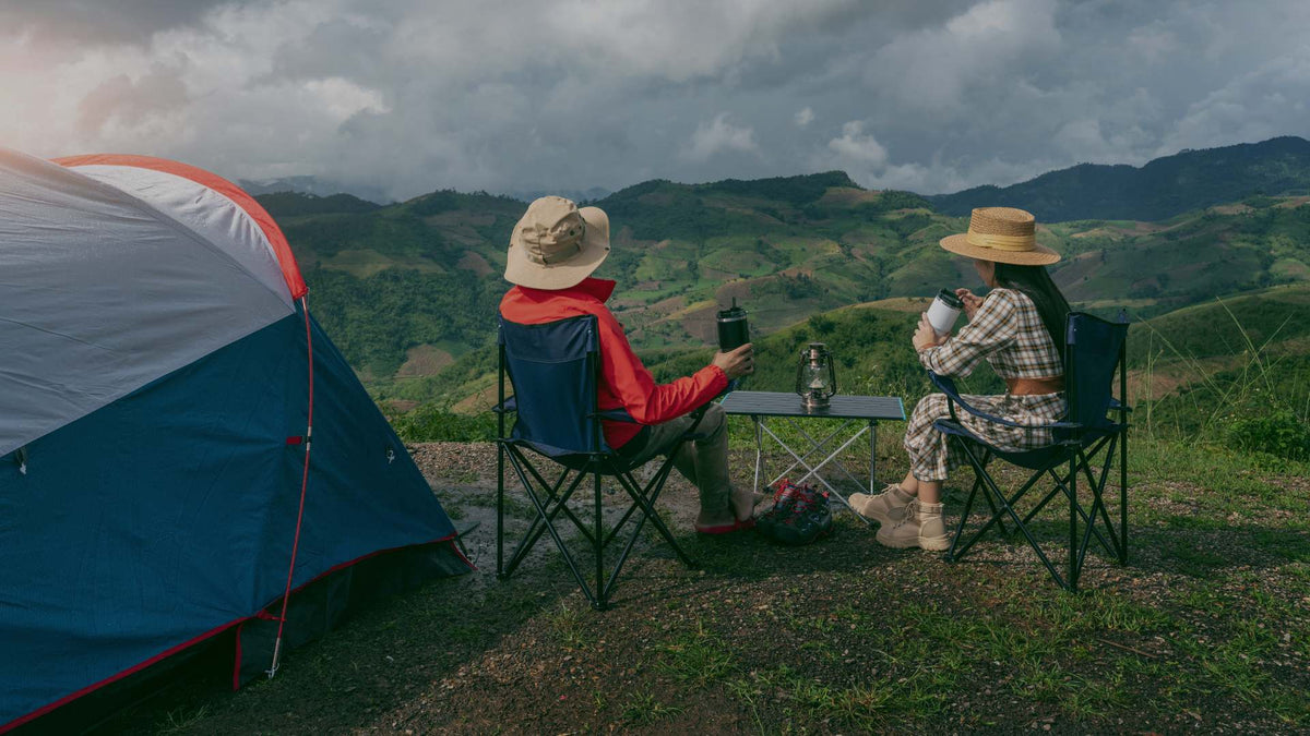 Two people sitting in camping chairs at a scenic mountain campsite under a cloudy sky, with a blue and white tent on the left and a small folding table between them.