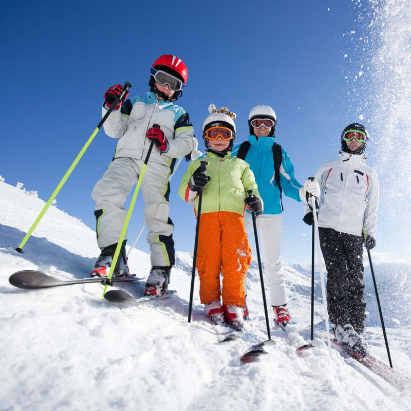 Four children in colorful ski suits and helmets pose on a snowy mountain slope with skis and ski poles under a bright blue sky.