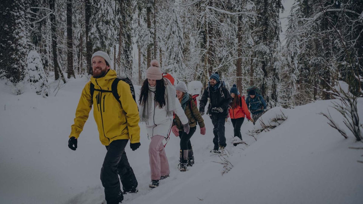 A group of people hiking through deep snow in a forest, with the most prominent individual in the foreground wearing a bright yellow jacket, black pants, and a white beanie.