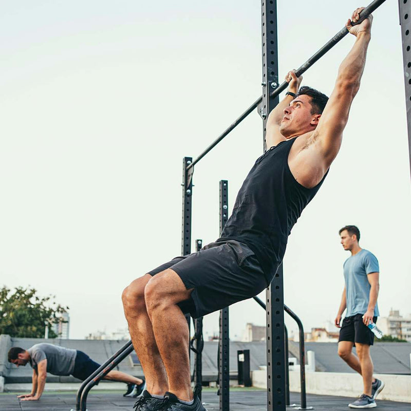 A man in a black tank top and shorts hangs from a pull-up bar on an outdoor rooftop gym, with other gym-goers visible in the background.