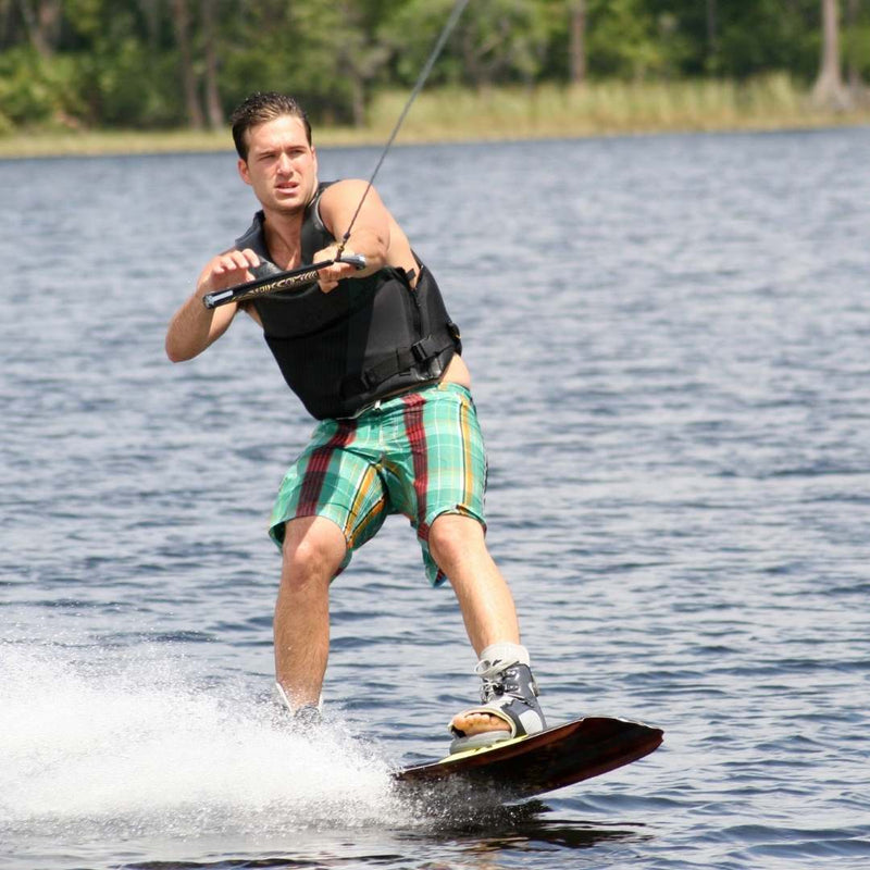 A man wakeboarding on a lake, wearing a black life vest, green and red plaid shorts, and holding a tow rope. A wakeboard is visible beneath his feet, creating a splash of water.