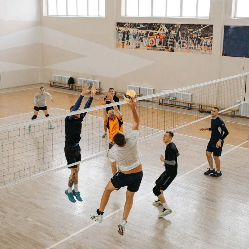 A group of men playing volleyball in a large indoor sports hall with wooden floors and high windows. Two players jump at the net to block the ball, while others position themselves on the court.