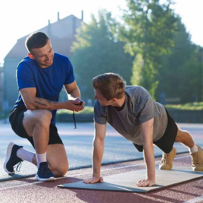 Two men outdoors on a running track, one kneeling and holding a stopwatch while the other performs a push-up on a yoga mat. The man in the foreground wears a gray t-shirt and black shorts, and the man behind wears a blue t-shirt and black shorts.