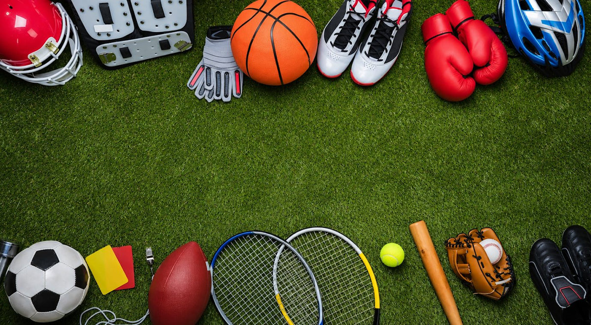 A flat-lay arrangement of various sports equipment on a green turf background, featuring a red football helmet, white cleats, red boxing gloves, a blue bicycle helmet, a basketball, a soccer ball, a football, tennis rackets, a bat, a baseball glove, and other athletic accessories.